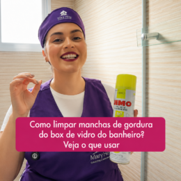 Mulher usando uniforme roxo da Mary Help segura um produto para limpeza de box e faz um gesto de aprovação com a mão, em um banheiro com box de vidro. Sobre a imagem, há uma caixa rosa com o texto “Como limpar manchas de gordura do box de vidro do banheiro? Veja o que usar”.